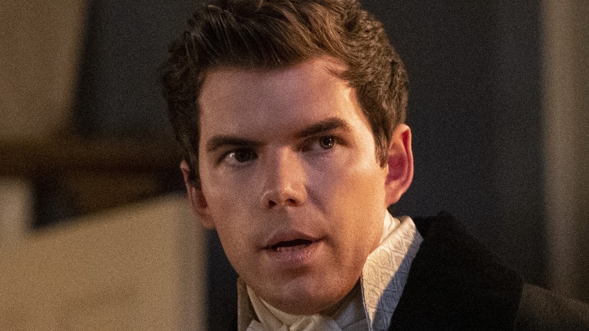 Close-up of Luke Newton as Colin on Netflix's 'Bridgerton.' He is a young, white man with short brown hair wearing a high-collared, Regency-era style shirt under a black jacket. His gaze is looking off to the side. 