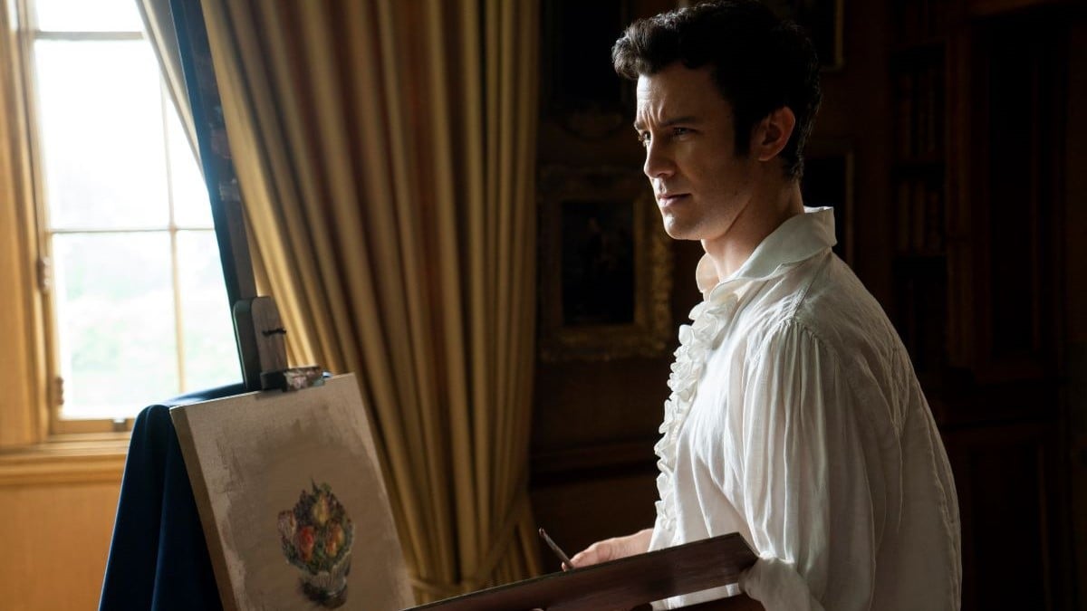 Image of Luke Thompson as Benedict in Netflix's 'Bridgerton.' He is a young, white man with dark hair and sideburns wearing a long-sleeved, frilly Regency-era shirt and holding a paint palate and standing in front of an easel where he's painting a still-life of a bowl of fruit. There is a window behind him letting in the daylight.