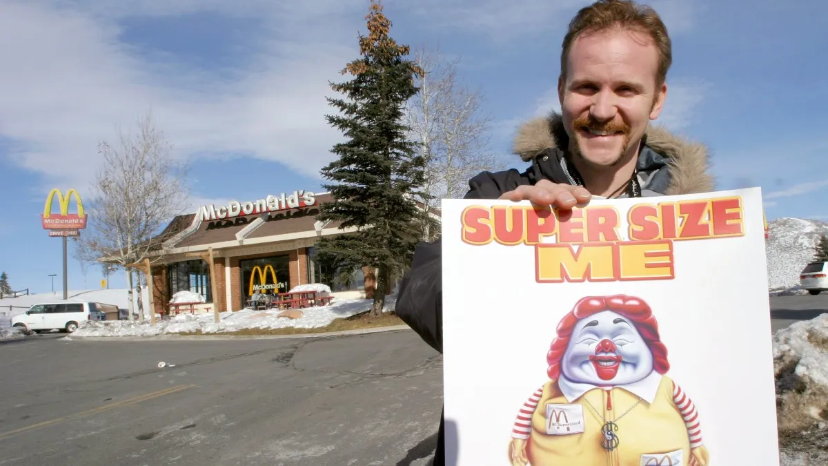 Morgan Spurlock posing outside of a McDonald's with a Super Size Me poster