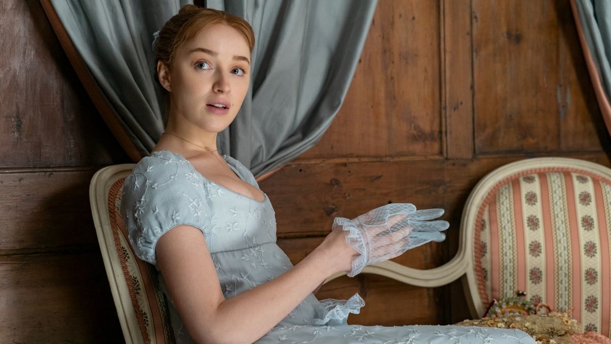 Image of Phoebe Dynevor as Daphne on Netflix's 'Bridgerton.' She is a young, white woman with red hair pulled up in a bun and wearing a light blue, Regency-era dress with cap sleeves and putting on sheer lace gloves. She's seated in a chair in a room with wood-paneled walls and talking to someone on her right. 