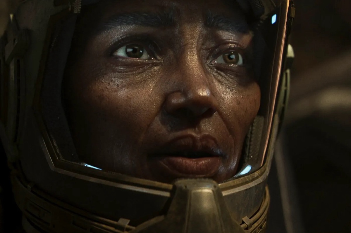 A woman looks pensively from behind a glass helmet in "Exodus Odyssey"