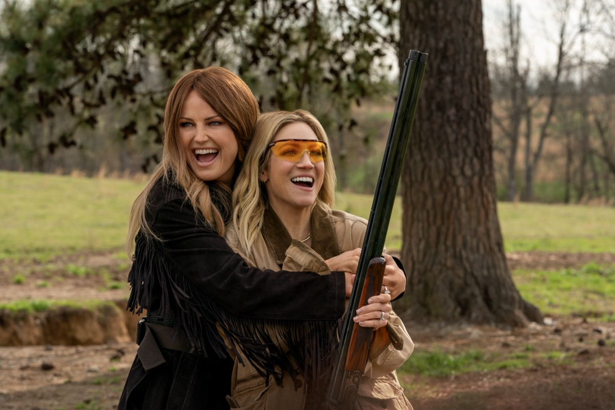Two women laugh and hug while holding a shotgun in "The Hunting Wives"