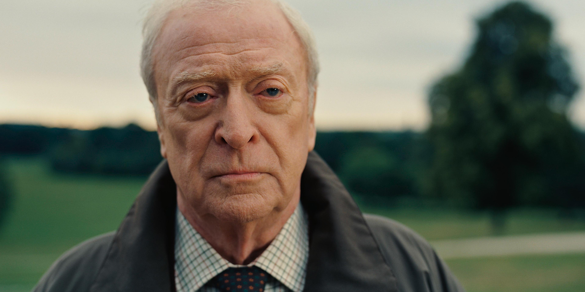 An old man wearing a grey coat and white shirt with pattern standing in a field.