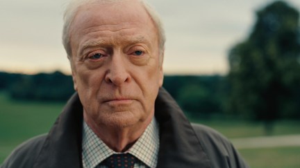 An old man wearing a grey coat and white shirt with pattern standing in a field.