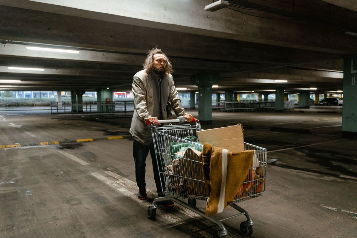A homeless man pushes a cart full of stuff in a parking garage.