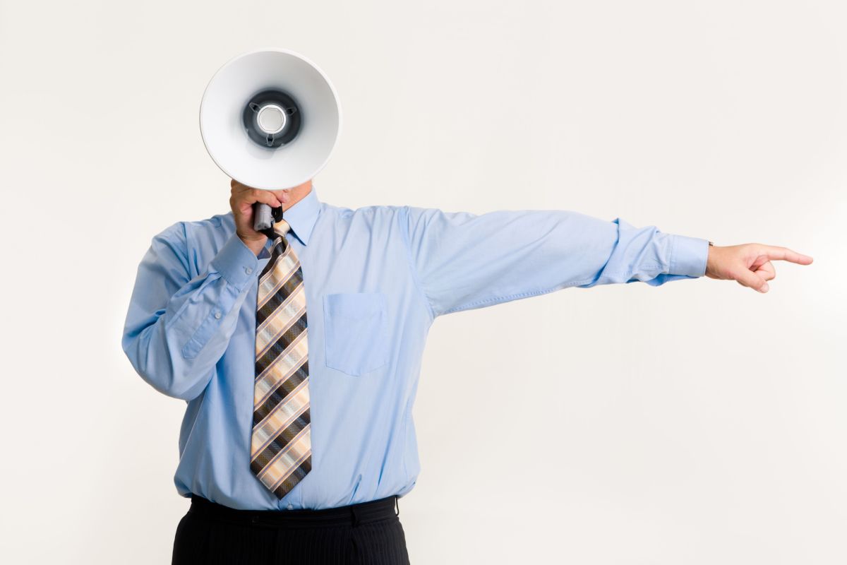 A man wearing business attire holds a megaphone up with his face covered while he points to the side with his left hand.