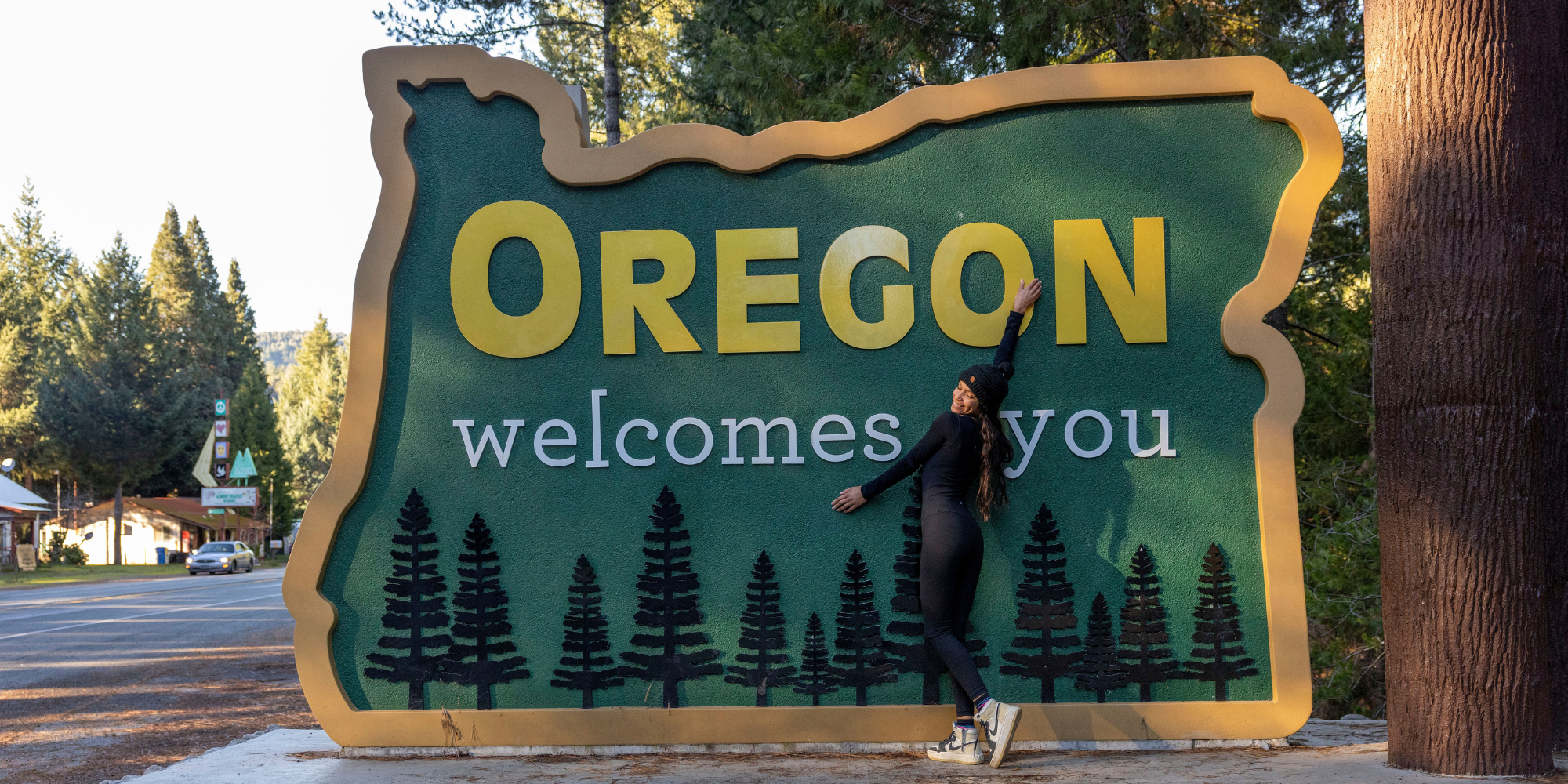 A woman stands in front of a sign reading "Oregon Welcomes You"