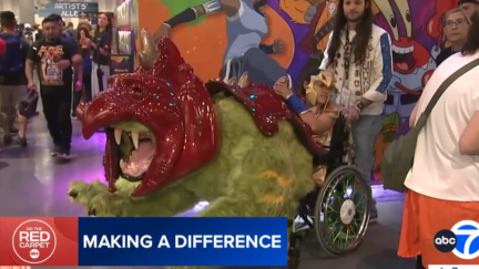 Screengrab from an interview at San Diego Comic Con featuring a girl in a cosplay wheelchair of Battle Cat from 
